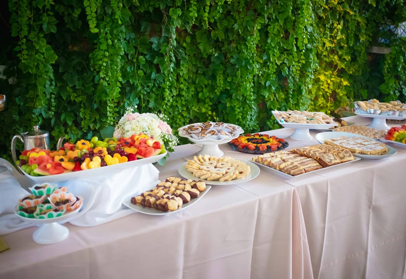 Buffet table with fruit platters, biscuits and traybakes on pink cloth against an ivy-covered wall.