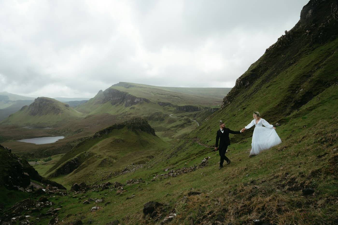 A bride and groom hold hands on a green hillside above a valley and loch at the Quiraing, Skye.